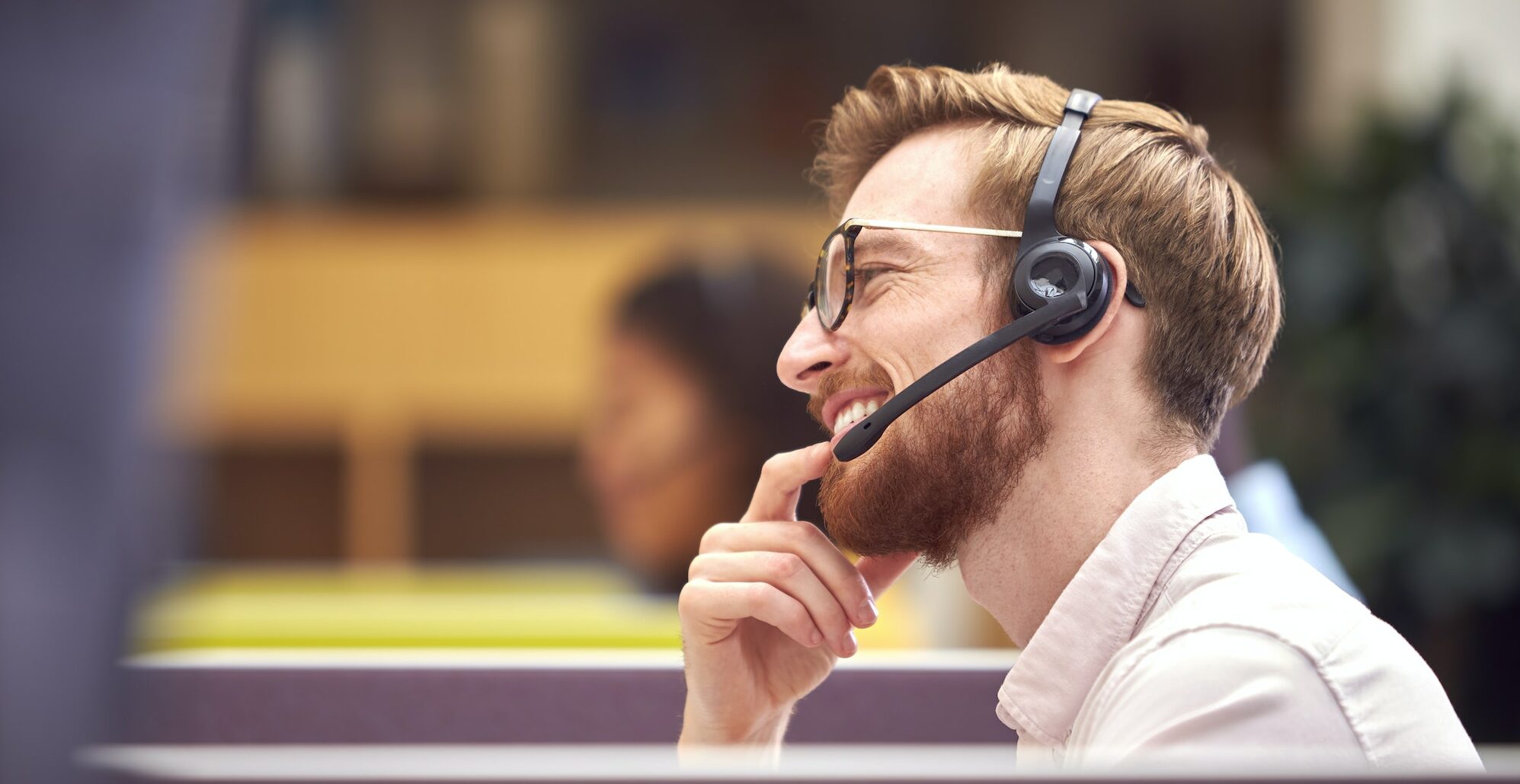 Businessman In Cubicle Wearing Headset Talking To Caller In Busy Customer Services Centre